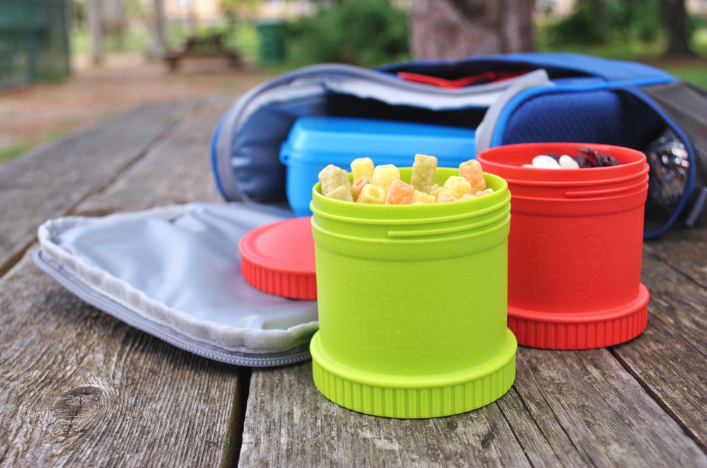 Envases Re-Play verde y rojo con snacks junto a una lonchera, sobre mesa de picnic